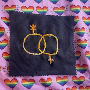 May include: A black fabric square with an embroidered symbol of two interlocking circles, one with a cross and the other with a star. The embroidery is in shades of yellow and orange. The background is a pink fabric with rainbow heart patterns.