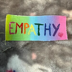 Może przedstawiać: Prostokątna naszywka z napisem "EMPATHY" w tęczowych literach. Tło naszywki przechodzi od zielonego do różowego. Małe różowe serce znajduje się obok litery "Y".