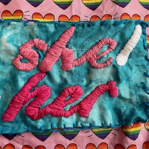 Op de afbeelding: Een turquoise stoffen lap met de woorden "she/her" geborduurd in roze draad. De lap is met blauwe draad langs de randen gestikt. De achtergrond is een roze stof met regenboog hart patronen.