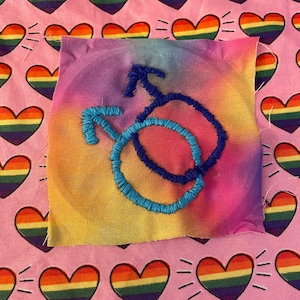 Puede incluir: Un parche de tela con un símbolo masculino bordado en azul sobre un fondo degradado arcoíris. El parche está colocado sobre una tela rosa estampada con corazones de colores del arcoíris.