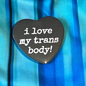 May include: A black heart-shaped pin with the white text "i love my trans body!" against a blue and teal striped background. The pin is a solid color with a glossy finish.