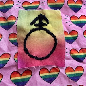 May include: A small, square fabric patch with a gradient of yellow, orange, and pink. A black embroidered symbol, resembling a circle with an arrow, is centered on the patch. The background is a pink fabric patterned with rainbow-colored hearts.