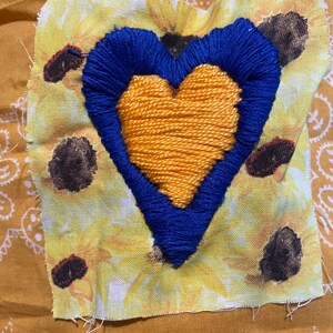 Puede incluir: Un parche de tela con un diseño de corazón. El corazón está bordado con hilo amarillo en el centro, rodeado por un contorno bordado azul. El parche está sobre una tela estampada con girasoles amarillos.
