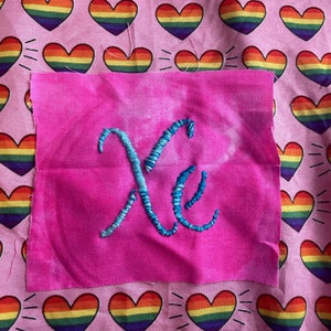 Op de afbeelding: Een roze stoffen vierkant met de geborduurde letters "Xe" in turquoise draad, tegen een achtergrond van roze stof met een patroon van regenboogkleurige harten. De harten hebben een regenboogverloop en witte lijnen die ervan uitstralen.