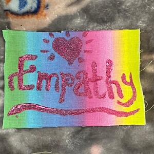 May include: A rectangular fabric patch with a rainbow gradient background. The word "Empathy" is written in glittery pink letters, with a glitter heart and sunburst design above the word. The patch is on a gray, textured surface.