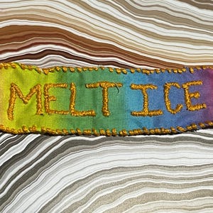 Può includere: Un pezzo di tessuto rettangolare, cucito a mano, con le parole "MELT ICE" ricamate con filo dorato. Il tessuto presenta una sfumatura di colori, che va dal rosa al giallo, verde e blu. I bordi sono delineati con cuciture dorate.