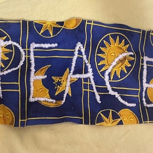 May include: A blue fabric featuring a celestial design of suns, moons, and stars. The word "PEACE" is embroidered in white thread. The fabric is rectangular with a slightly frayed edge, creating a unique aesthetic.