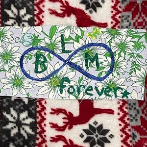 May include: A fabric patch with the text "BLM forever" in green and blue, inside an infinity symbol. The patch is set against a floral background with white flowers and green leaves. The background is surrounded by a red and white patterned fabric.