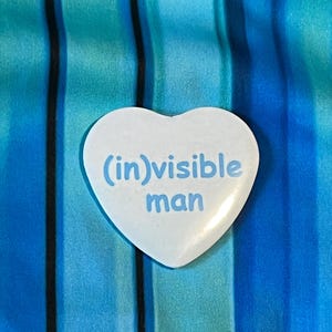 Puede incluir: Un botón blanco en forma de corazón con las palabras "(in)visible man" en letras azul claro. El botón está sobre un fondo de tela a rayas azules y turquesas.