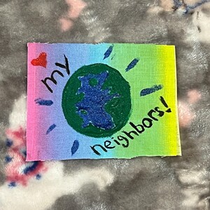Può includere: Un'opera d'arte rettangolare dipinta a mano con uno sfondo sfumato arcobaleno. Presenta un cerchio verde con un centro blu, le parole "my neighbors!" e un cuore rosso.