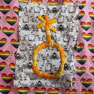 Op de afbeelding: Een geborduurd symbool in tinten geel en oranje, gestikt op een stuk stof met een zwart-wit patroon. De stof is geplaatst tegen een roze achtergrond versierd met regenboog hartvormen.