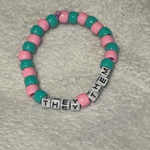 May include: A beaded bracelet with pink, teal, and white beads. The bracelet has the word "THEY" spelled out in white letter beads. The bracelet is made of round beads strung together on a stretchy cord.