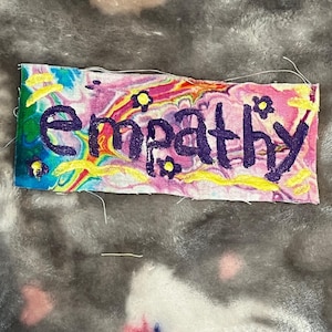 Pode incluir: Um patch de tecido retangular com a palavra "empathy" bordada em roxo. O fundo apresenta um design abstrato colorido com redemoinhos de rosa, azul, verde e amarelo. Pequenas flores amarelas estão espalhadas ao redor do texto.