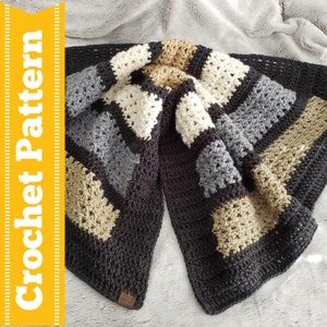 May include: A crocheted blanket with a striped pattern in black, beige, grey, and white. The blanket is folded in half and has a small brown leather tag with the text "The Winter Collection" on it.