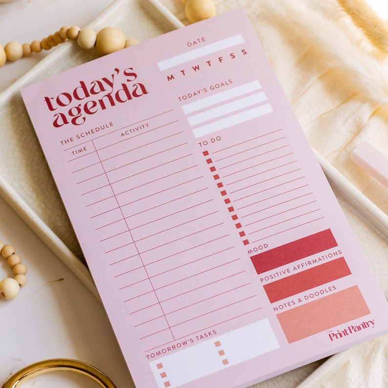 K&ouml;nnte beinhalten: Ein rosa Tagesplaner mit Abschnitten f&uuml;r den Zeitplan, die To-Do-Liste, die Stimmung, positive Affirmationen, Notizen und Kritzeleien sowie Aufgaben f&uuml;r morgen. Der Planer hat einen roten und wei&szlig;en Titel, der "today's agenda" lautet.