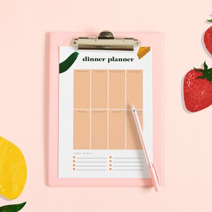 May include: A pink clipboard with a white sheet of paper that says "dinner planner" and has a weekly meal planner grid. The clipboard has a silver clip and a pen is next to it. There are two red strawberries and two yellow lemons on a pink background.