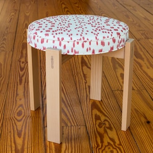 May include: A small, round stool with a light wood frame and a patterned seat. The seat cushion is white with a repeating pattern of red brushstrokes. The stool has four straight legs.