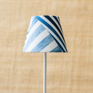 May include: A small lampshade with a white base and a blue and white striped pattern. The lampshade is a trapezoid shape and is mounted on a white pole. The background is a light brown color.
