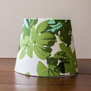 May include: A tapered drum lampshade featuring a white background with a repeating pattern of large, stylized fig leaves in various shades of forest green, lime green, and light mint green, creating a bold botanical print across the surface.