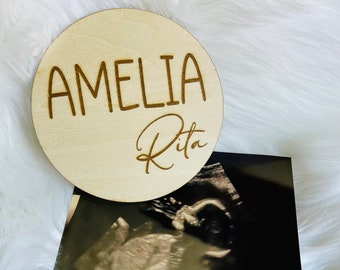 Puede incluir: Una placa redonda de madera con el nombre "Amelia Rita" grabado en escritura cursiva. La placa está encima de una imagen de ultrasonido en blanco y negro.