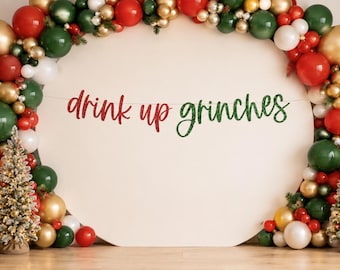 May include: A circular backdrop featuring a balloon arch in red, green, white, and gold. A banner with the text "drink up grinches" in red and green script hangs across the center. Four small, flocked artificial Christmas trees stand in front.