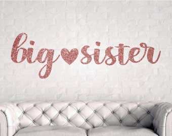 Puede incluir: Pancarta de purpurina dorada rosa con el texto "big heart sister" colgada en una pared de ladrillo blanco.