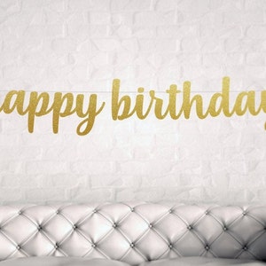 May include: Gold glitter happy birthday banner hanging on a white brick wall.