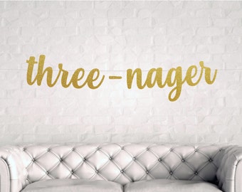 May include: Gold glitter banner with the text "three-nager" hanging on a white brick wall.