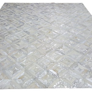 May include: A white and silver cowhide rug with a geometric pattern. The rug is made of individual pieces of cowhide that have been sewn together to create a unique design.