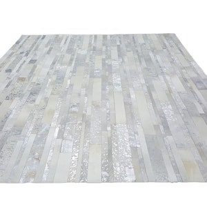 May include: A rectangular rug with a patchwork design. The rug features a mix of light grey, white, and silver rectangular pieces arranged in a linear pattern. The rug has a textured appearance and a neutral colour palette.