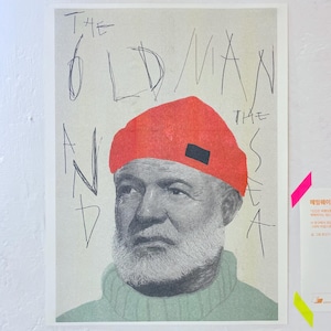 以下が含まれることがあります： 赤い帽子をかぶった老人の白黒イラストが描かれたポスター。ポスターには黒いマーカーで「THE OLD MAN AND THE SEA」という文字が書かれている。