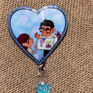 Könnte beinhalten: Ein herzförmiges Ausweis-Jojo mit einer Cartoon-Illustration eines Mannes und einer Frau. Das Bild enthält den Text "Carl & Ellie". Das Ausweis-Jojo hat einen transparenten Kunststoffclip mit einem blauen Glitzerherz.