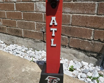 Puede incluir: Un abridor de botellas de madera roja con un abridor de botellas de metal negro adjunto. El abridor de botellas tiene las letras ATL en blanco en la parte delantera. La parte inferior del abridor de botellas es negra con un logotipo rojo de los Atlanta Falcons.