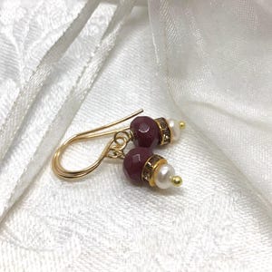 May include: Gold drop earrings with faceted burgundy beads, small pearls, and gold accents. The earrings have a delicate design with a hook closure. The jewellery is set against a white, textured background.