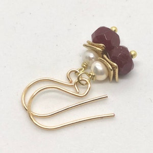 May include: Gold earrings featuring two burgundy faceted beads and two white pearls. The earrings have a simple hook fastening.