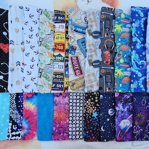May include: A collection of fabric bookmarks with various patterns. Designs include anchors, arrows, license plates, Route 66, camping themes, beach scenes, mermaids, stars, planets, and more. The bookmarks are rectangular and feature a variety of colors and styles.