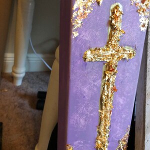 Könnte beinhalten: Ein violettes Leinwandbild mit einem goldenen Kreuz in der Mitte. Das Kreuz besteht aus Blattgold und hat eine strukturierte Oberfläche. Die Leinwand ist von einem Rand aus Blattgold umgeben.