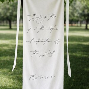 May include: Cream-coloured banner with black script. The text reads "Bring them up in the nurture and admonition of the Lord. Ephesians 6:4." The banner is hanging from a tree branch with white ribbons.