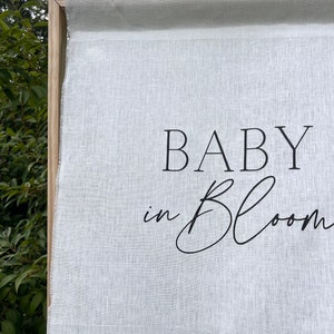 Può includere: Striscione in tessuto bianco con testo nero che recita "BABY in BLOOM".