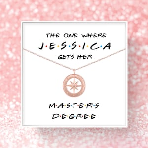 May include: A white card with the text "The One Where Jessica Gets Her Master's Degree" in a Friends-themed font. A rose gold necklace with a compass pendant is displayed on the card.
