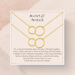 May include: A gold necklace set with two interlocking circles, presented in a white box with the text "AUNT & NIECE" and a message about the special bond between aunt and niece.
