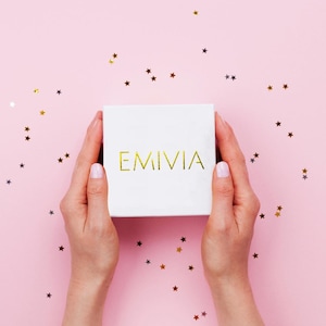 May include: A white gift box with the word "EMIVIA" printed in gold on a pink background with gold glitter stars.