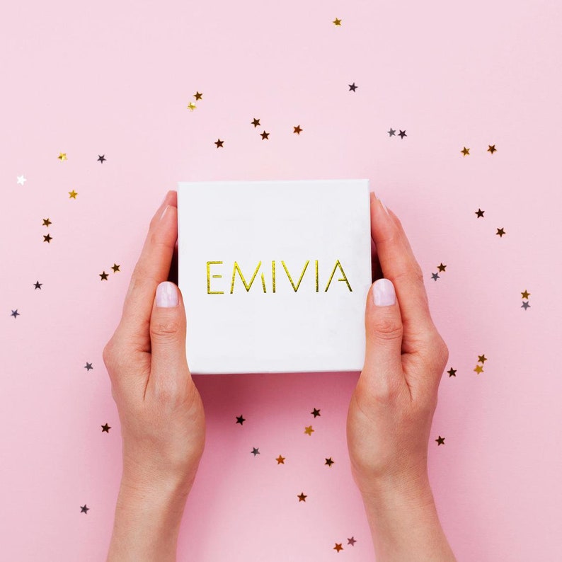 May include: A white gift box with the word "EMIVIA" printed in gold on a pink background with gold glitter stars.