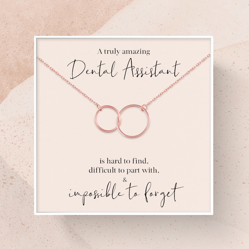 May include: A white card with a rose gold necklace featuring two interlocking circles. The card reads "A truly amazing Dental Assistant is hard to find, difficult to part with, & impossible to forget."