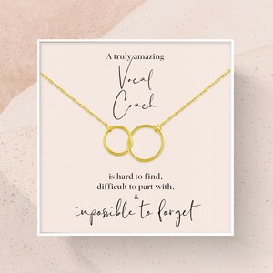 May include: A white card with gold lettering that reads "A truly amazing Vocal Coach is hard to find, difficult to part with, & impossible to forget". A gold chain necklace with two interlocking circles hangs from the top of the card.
