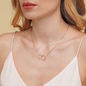 May include: A delicate rose gold necklace with two interlocking circles. The necklace is on a model wearing a white tank top.