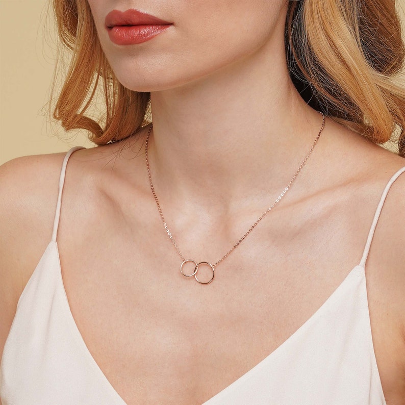 May include: A delicate rose gold necklace with two interlocking circles. The necklace is on a model wearing a white tank top.