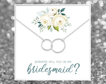 Peut inclure: Une carte blanche avec un motif floral et un collier argenté avec deux cercles entrelacés. La carte dit "Jennifer, veux-tu être ma demoiselle d'honneur ?"