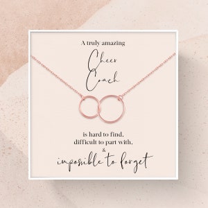 May include: A white card with a rose gold necklace featuring two interlocking circles. The card reads "A truly amazing Cheer Coach is hard to find, difficult to part with, & impossible to forget."