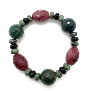 May include: A beaded bracelet featuring faceted round and oval beads in shades of deep red, forest green, and black. The stones have a polished, textured surface and are arranged in a repeating pattern around the circular elastic cord structure.